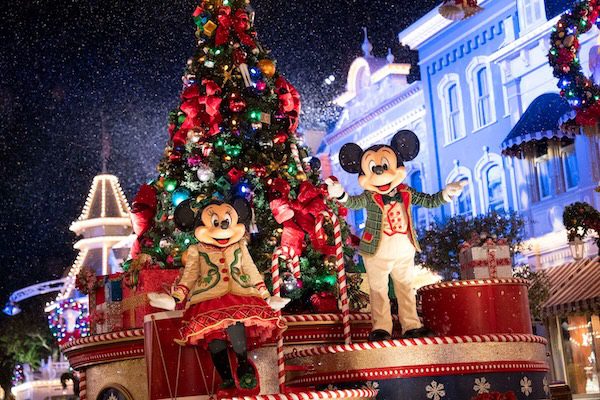 Mickey and Minnie Mouse joyfully pose in festive outfits beside a beautifully decorated Christmas tree, with snowy lights in the background.