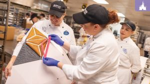 Pastry chefs at EPCOT assemble a large gingerbread replica of Spaceship Earth, carefully positioning handcrafted gingerbread pieces in a kitchen workspace.