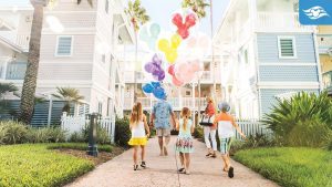Family walking toward pastel-colored Disney resort buildings, carrying colorful Mickey-shaped balloons.