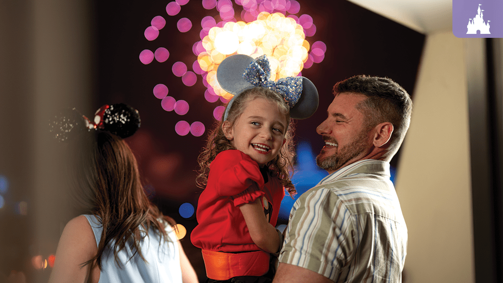 Smiling father holding daughter in Mickey ears while watching colorful fireworks at Walt Disney World Resort.