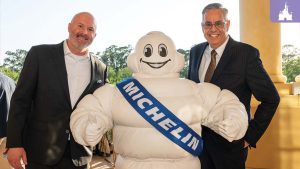 Two men in suits stand smiling on either side of the Michelin Man mascot wearing a blue sash, celebrating Victoria & Albert’s MICHELIN Star award.