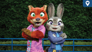 Nick Wilde and Judy Hopps pose back-to-back in their Zootopia 2 outfits at a Disney park, smiling in front of lush greenery.