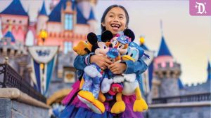 Smiling child in front of Sleeping Beauty Castle holding plush toys of Mickey, Minnie, Donald, and Goofy in Disneyland 70th Anniversary outfits.
