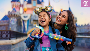 Smiling child wearing golden Mickey ears and holding a glowing 70th anniversary wand at Disneyland, with Sleeping Beauty Castle in the background.