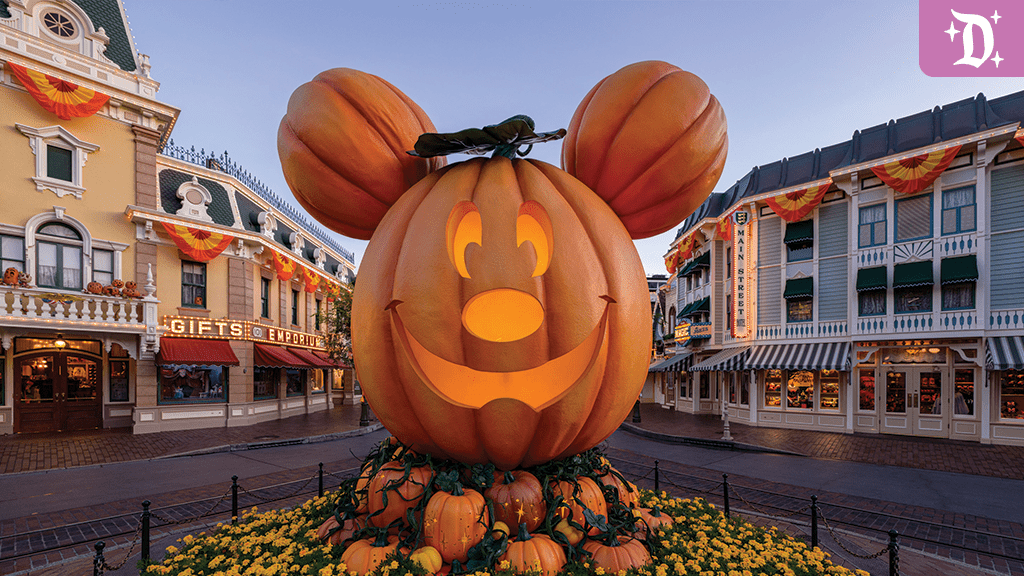 A large smiling Mickey Mouse-shaped pumpkin sits at the center, surrounded by smaller pumpkins, with festive decorations in a lively street.