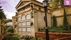 Stone mausoleum with ornate epitaphs, surrounded by lush greenery and brick accents, located in the expanded grounds of Disneyland's Haunted Mansion.