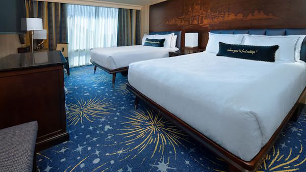 A hotel room with two queen beds, a colorful starry carpet, and a large window letting in natural light. Decor features a city skyline.