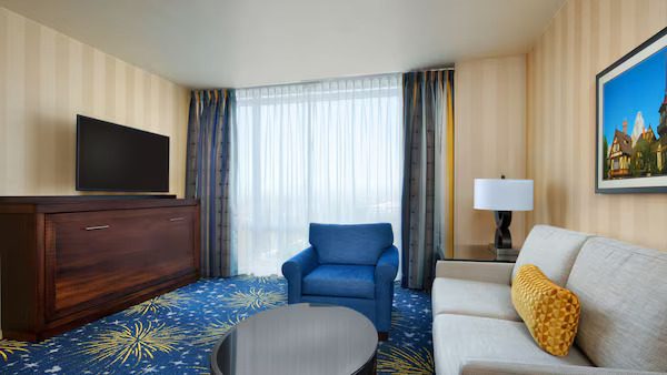 A modern hotel room featuring a sofa, blue armchair, a television, and a lamp, with large windows allowing natural light.