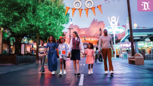 A family walks with a Disney VIP tour guide through a festively decorated Cars Land at Disney California Adventure during Oogie Boogie Bash, with Halloween lights and ornaments glowing.