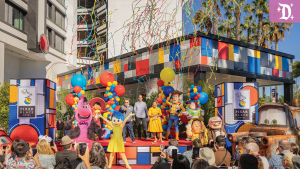 A colorful outdoor event features animated characters, balloons, and a crowd, with a backdrop of festive decorations and lively atmosphere.