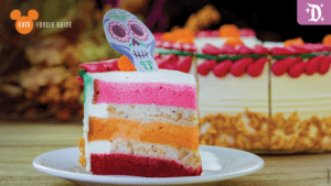 A vibrant slice of layered cake with pink, orange, and yellow stripes, topped with a colorful sugar skull decoration on a white plate.