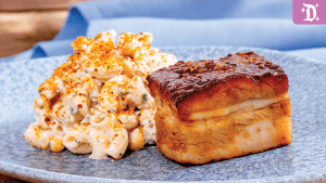 A plate featuring a piece of glazed pork belly next to a serving of creamy macaroni and cheese, garnished with crispy breadcrumbs.