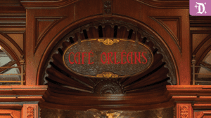 A wooden sign displaying "CAFÉ ORLEANS" in red text, set against an ornate background with intricate architectural details.