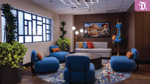 A cozy lounge area featuring blue chairs, a gray sofa, colorful pillows, and decorative plants, with vibrant artwork on the wall.