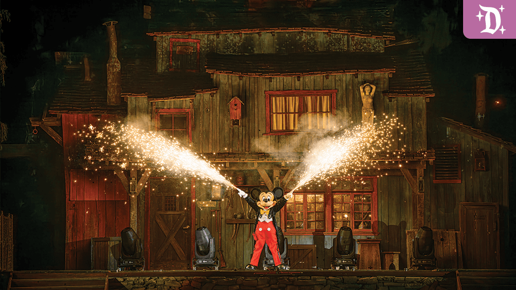 Mickey Mouse stands on stage in front of a rustic cabin, shooting sparks from handheld devices, surrounded by dramatic lighting.
