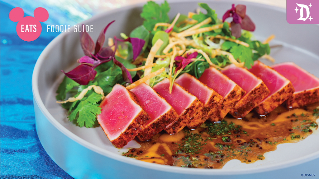 disneyland-resort-hotel-palm-breeze-foodie A plate featuring sliced seared tuna, drizzled with sauce, accompanied by fresh greens and colorful garnishes on a blue background.
