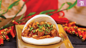 A fluffy bao bun filled with savory chicken, garnished with green onions, served on a golden plate with festive decorations in the background.