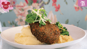 A plate featuring a brown, breaded patty on creamy mashed potatoes, garnished with greens and accompanied by steamed broccoli.