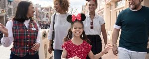 A joyful girl in a red dress and Minnie Mouse ears walks with family and a staff member at a theme park, all smiling and engaged.