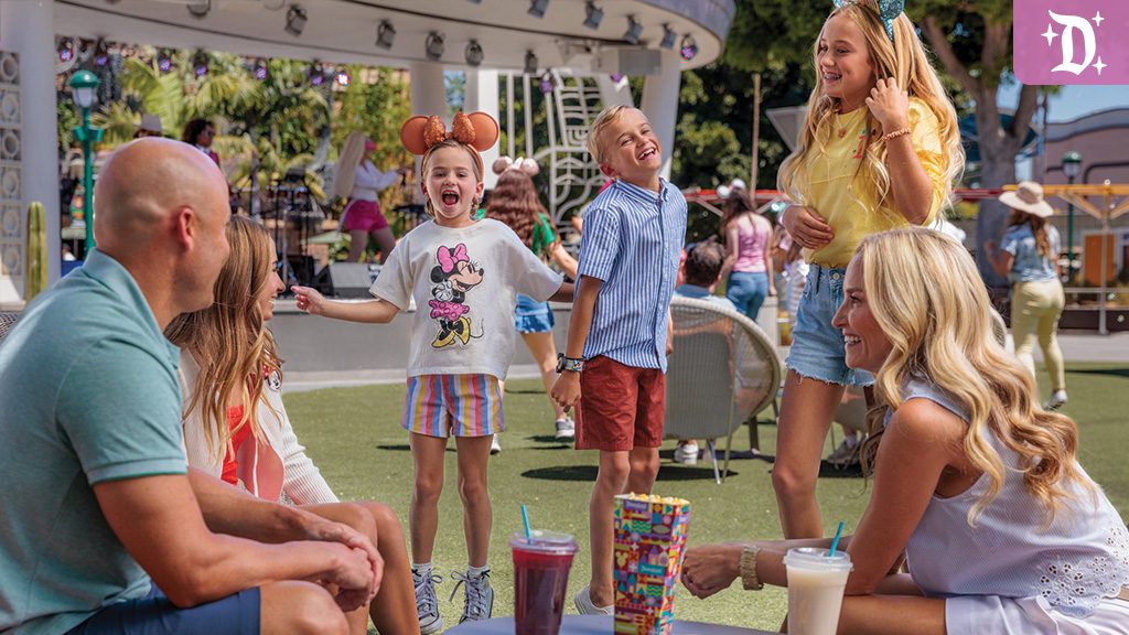 A family enjoys a sunny day at Downtown Disney District, with kids laughing in Disney-themed attire and drinks on the table.