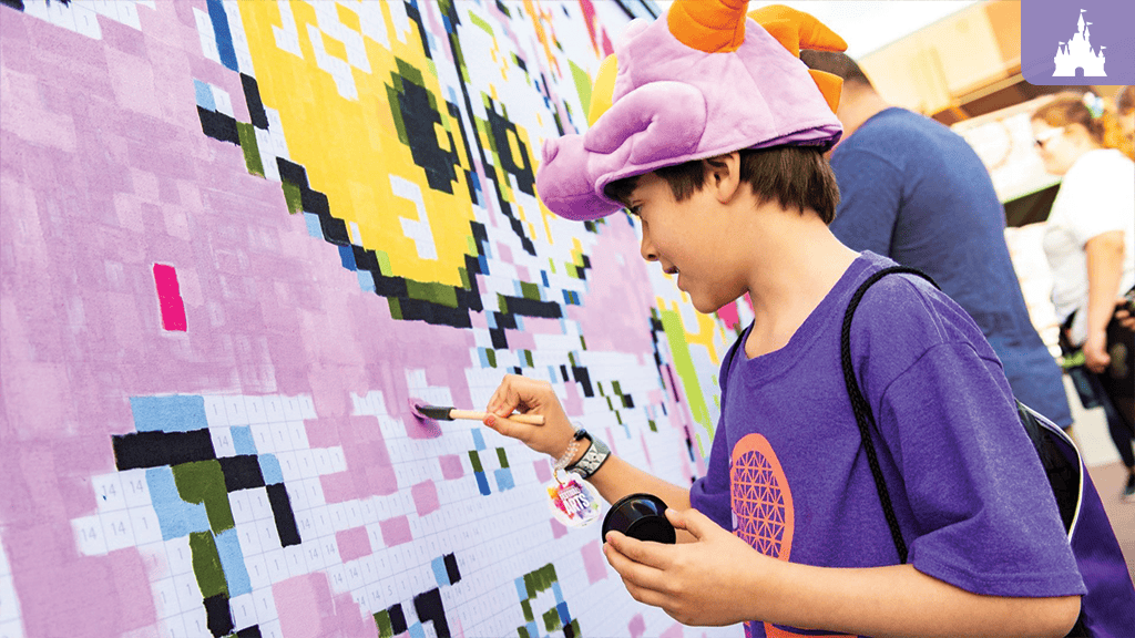 Child in purple shirt and dragon hat paints numbered squares on a large outdoor pixel-art mural with a brush and paint palette.