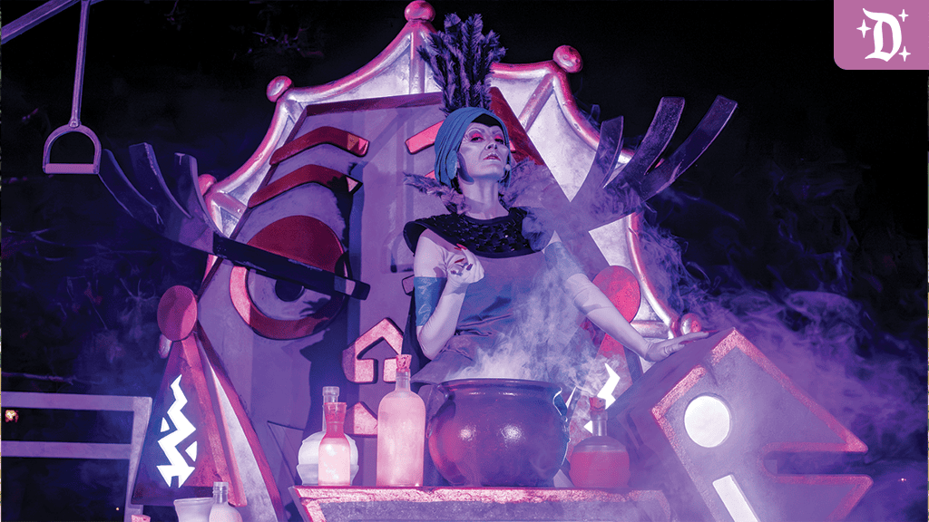 Performer in theatrical costume stands behind a smoking cauldron on stage decorated with a large stylized mask and glowing potion bottles