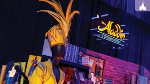 A vibrant costume from Aladdin on Broadway displayed on a mannequin, featuring an ornate gold and yellow headpiece with feather details. The background showcases the "Aladdin Costume Studio" sign and various costume design materials.