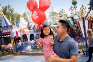 A father holds his smiling daughter in a theme park, both enjoying a red Mickey Mouse balloon amidst colorful attractions.