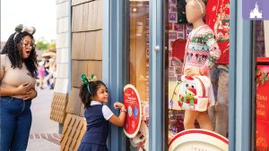 A young girl with festive Minnie Mouse ears interacts with a holiday-themed World of Disney store window using her MagicBand+, while an adult beside her looks on with delight.
