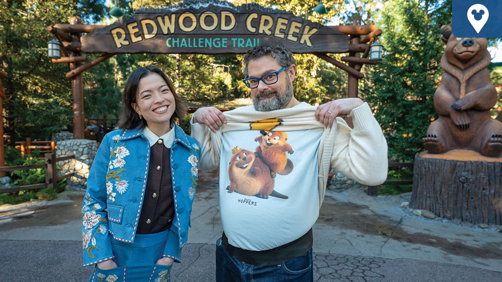 Two guests pose under the Redwood Creek Challenge Trail sign at Disney California Adventure, showing a Pixar Hoppers character T-shirt.