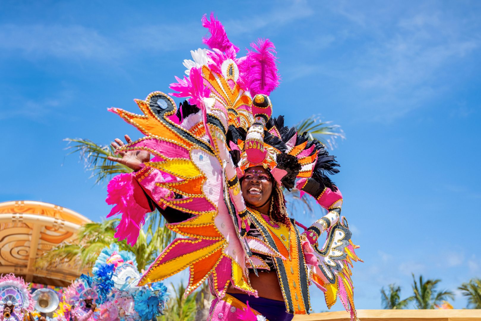 Vibrant Junkanoo performer in a bright, feathered costume dancing joyfully during a lively parade with a blue sky backdrop.