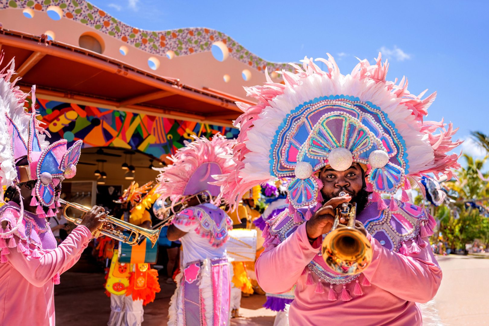 Colorful Junkanoo-style brass band performing in festive costumes outside a vibrant building under a bright blue sky.