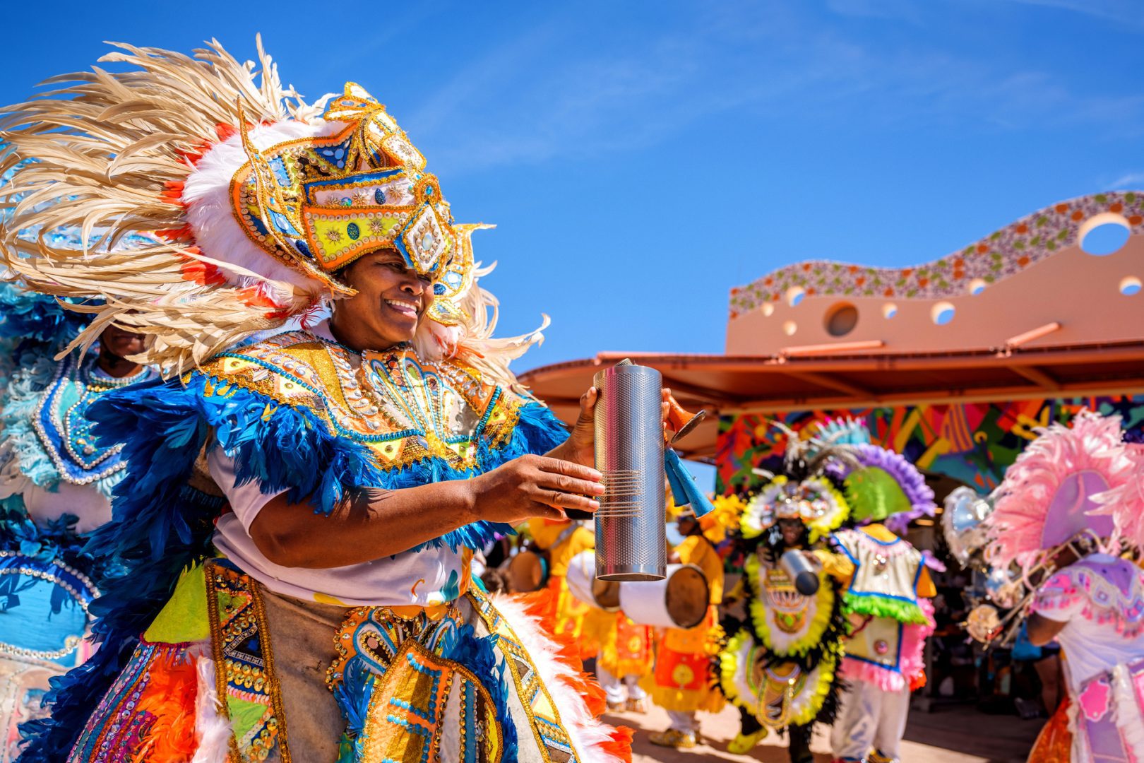 Smiling performer in vibrant Junkanoo costume with feathers and beads playing a metal percussion instrument at a festive outdoor event.
