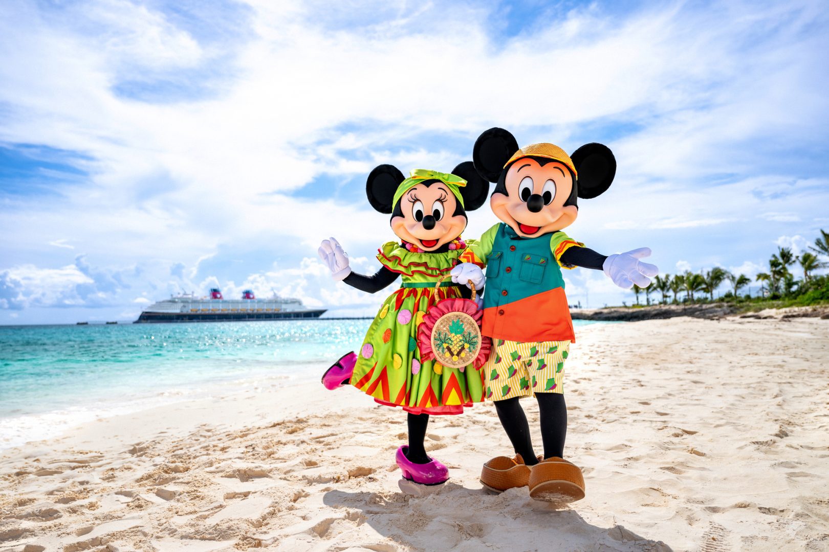 Mickey and Minnie Mouse in tropical outfits posing on a sunny beach with a Disney Cruise ship in the background.