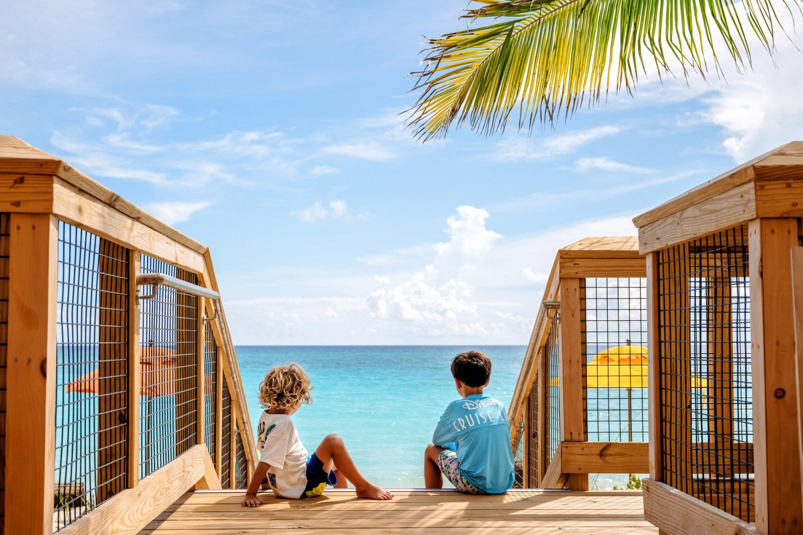 Two children sitting on a wooden platform, gazing out at the turquoise ocean under a bright sky with palm leaves framing the view.