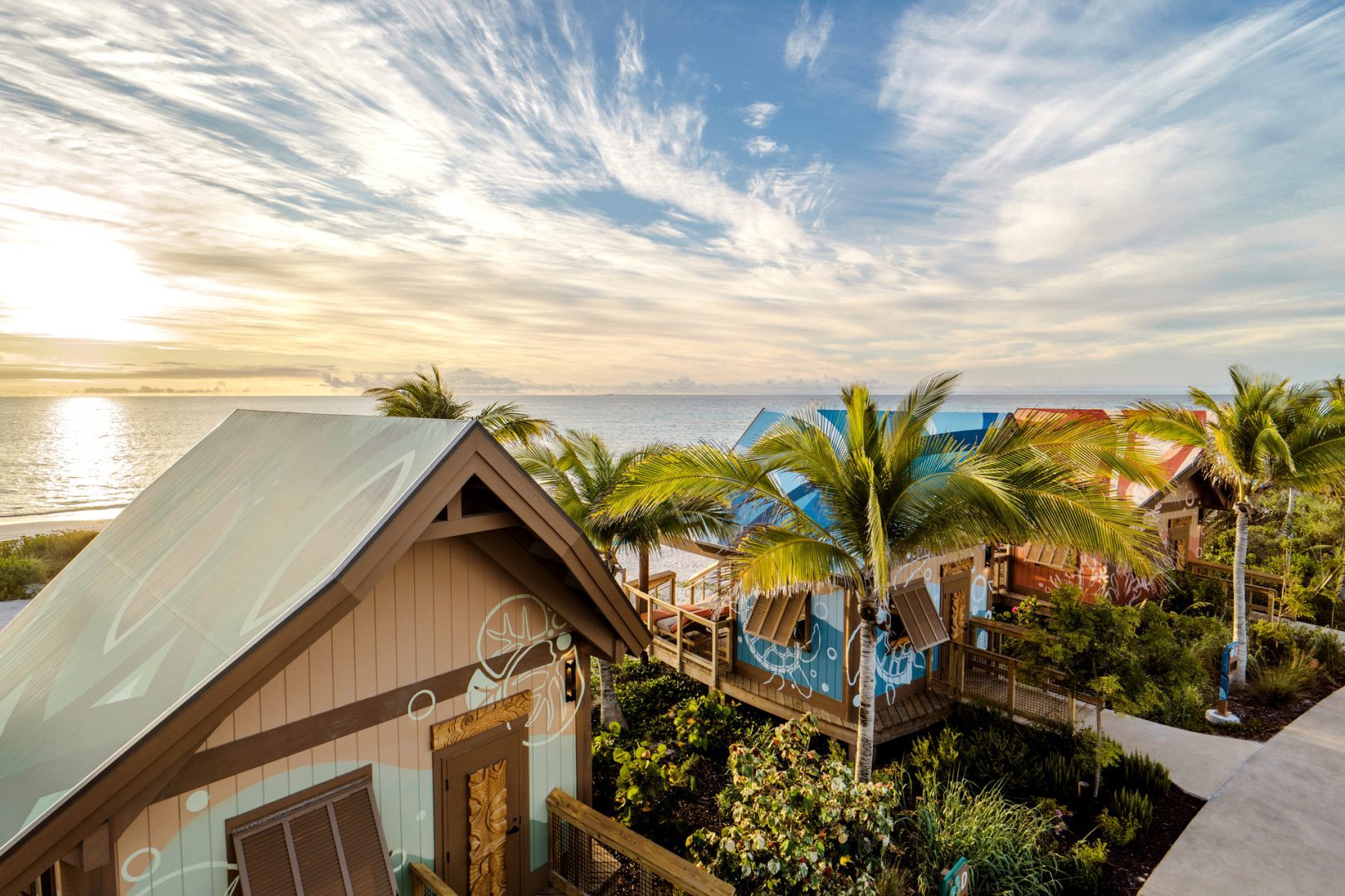 Cozy beachside cabanas with colorful roofs and ocean-inspired murals, surrounded by palm trees and overlooking a calm sea at sunset.