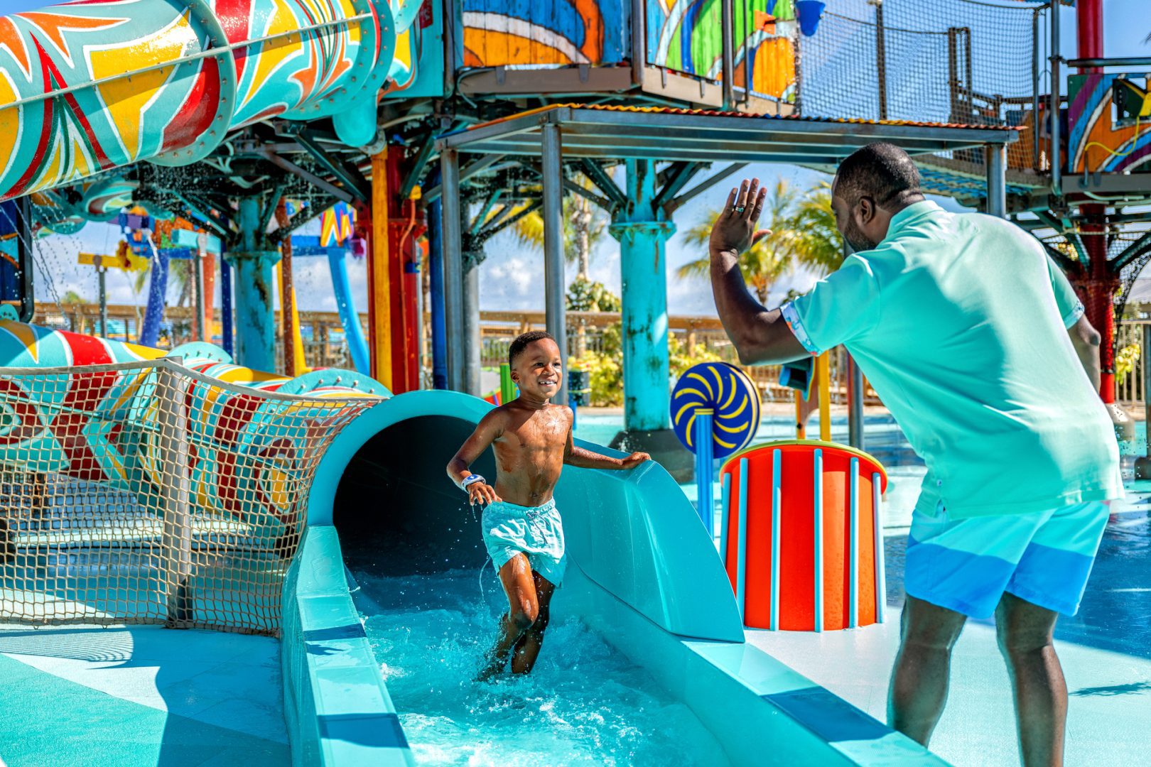 Young boy running out of a colorful water slide at a Disney-themed splash zone, smiling and high-fiving an adult.