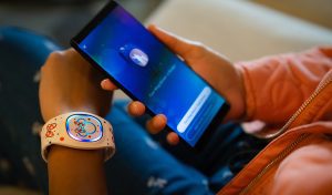 A child is holding a smartphone while wearing a colorful smartwatch. The background is softly blurred, suggesting a cozy indoor setting.