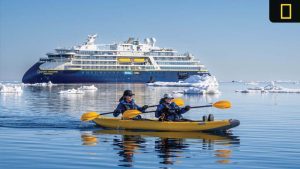 Two people kayak through icy Arctic waters with the National Geographic Endurance expedition ship in the background under clear blue skies.