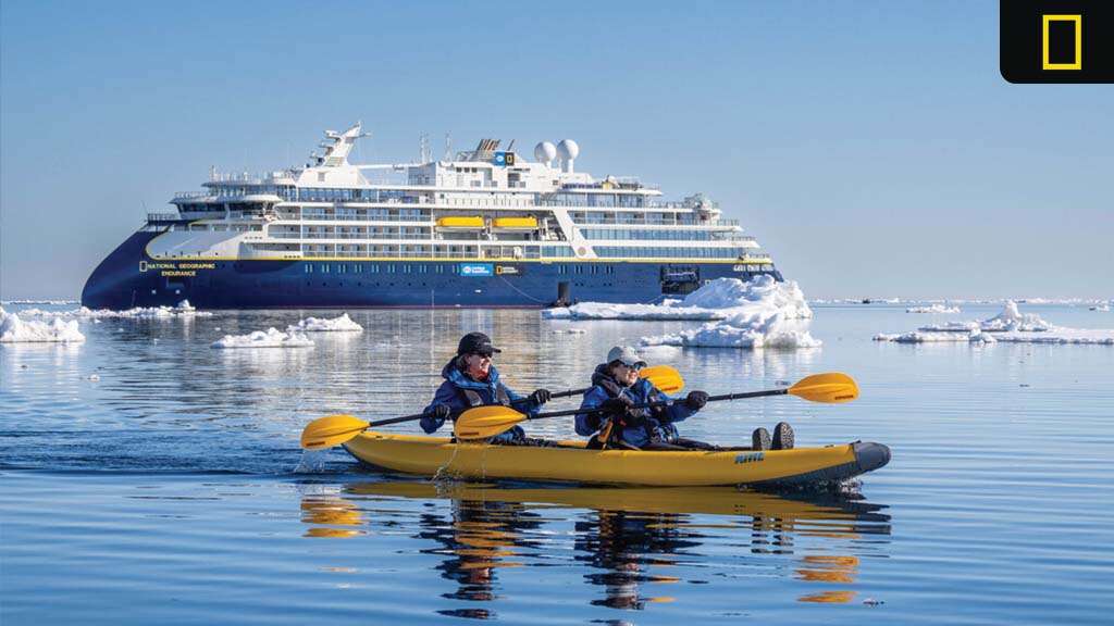 nat-geo-expeditions-2027-2028 Two people kayak through icy Arctic waters with the National Geographic Endurance expedition ship in the background under clear blue skies.