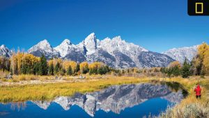 Snow-capped mountain peaks reflected in a calm lake, surrounded by autumn trees and a person in red taking a photo.