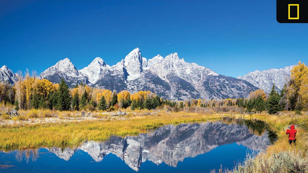 Snow-capped mountain peaks reflected in a calm lake, surrounded by autumn trees and a person in red taking a photo.