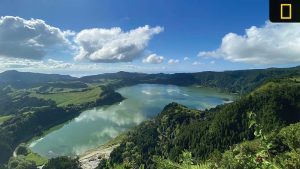 A panoramic view of a serene lake surrounded by lush greenery and rolling hills under a partly cloudy sky.