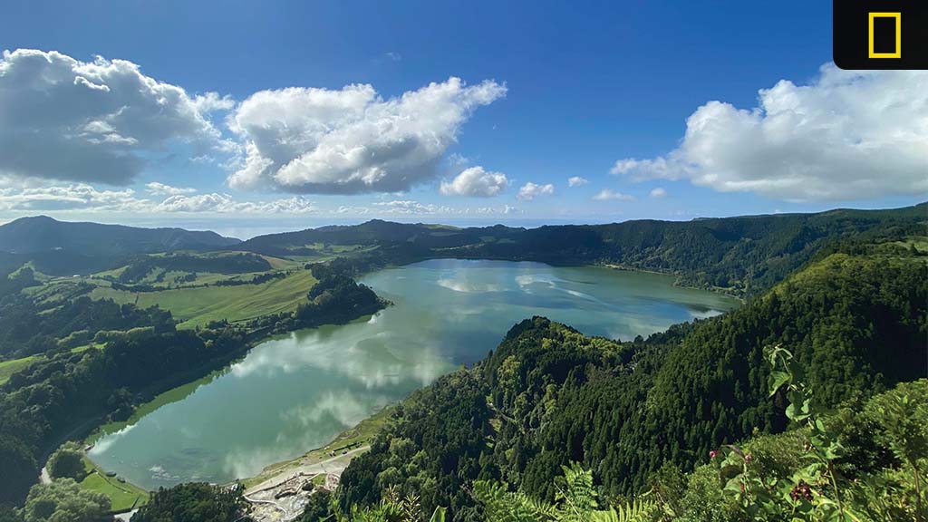 A panoramic view of a serene lake surrounded by lush greenery and rolling hills under a partly cloudy sky.