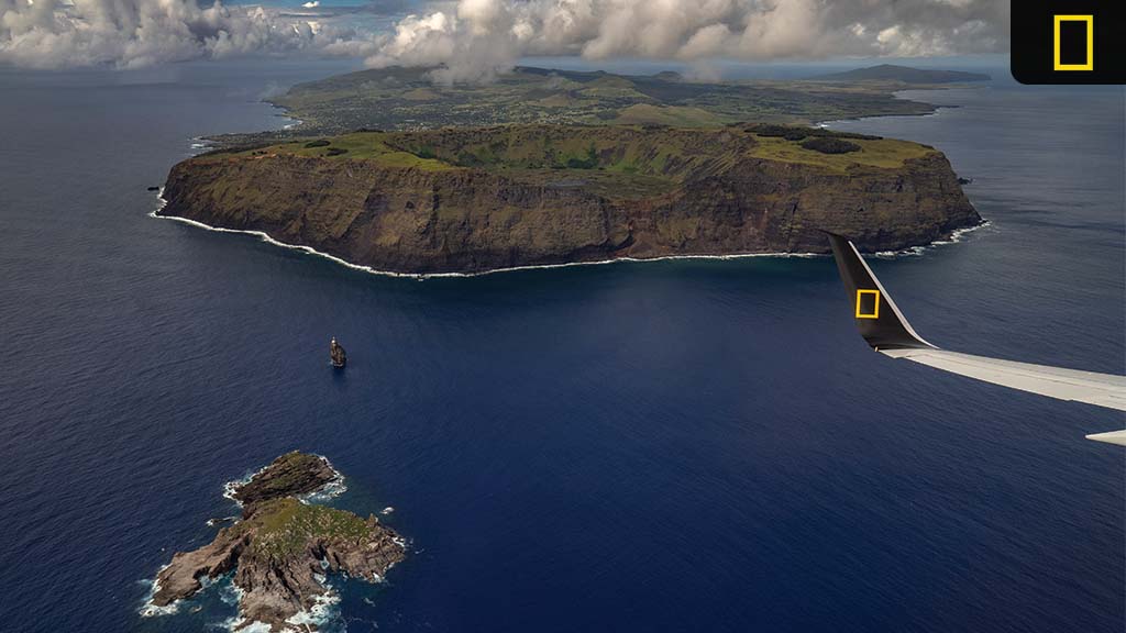 national-geographic-expeditions-around-the-world Aerial view of a large island and a smaller rocky outcrop, surrounded by deep blue ocean and dotted with clouds above.