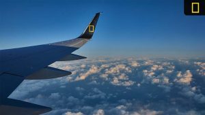 Aerial view of clouds from an airplane window, featuring the aircraft's wing with the National Geographic logo on the tail.