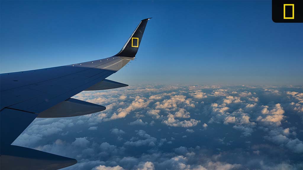 Aerial view of clouds from an airplane window, featuring the aircraft's wing with the National Geographic logo on the tail.