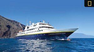 National Geographic Gemini expedition ship sailing near rocky Galápagos coastline under a clear blue sky.