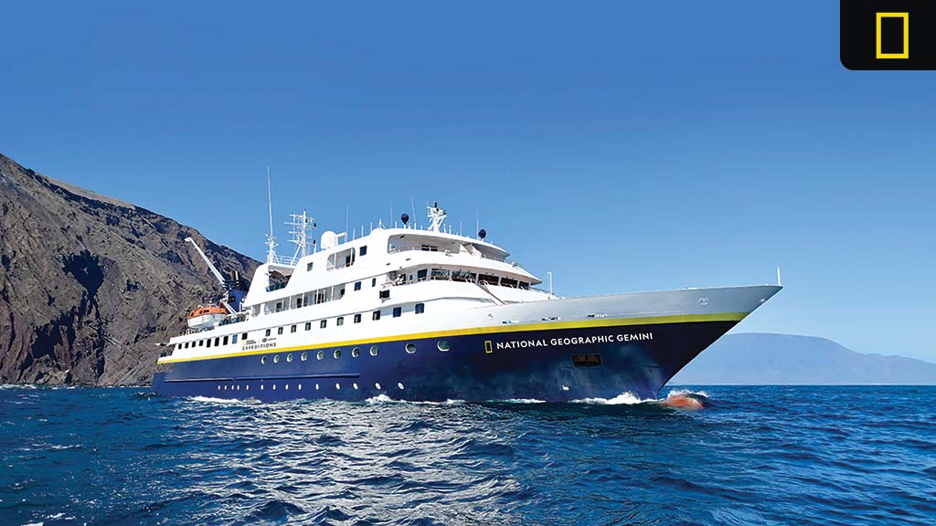 National Geographic Gemini expedition ship sailing near rocky Galápagos coastline under a clear blue sky.