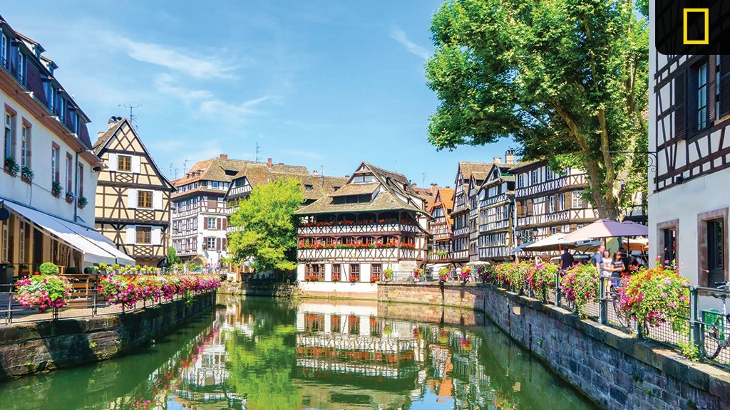 new-nat-geo-river-expeditions-2026 Half-timbered houses line a flower-decked canal in Strasbourg, France, under a clear blue sky.